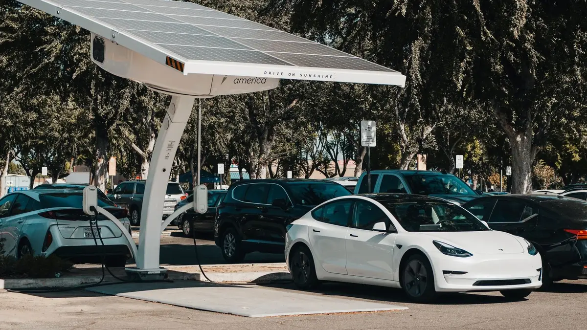 Une voiture électrique compacte branchée sur une borne de recharge, devant un parking urbain ensoleillé.